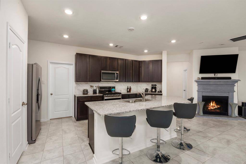 Modern kitchen with dark wood cabinets and stainless steel appliances. Features a large island with bar seating and a cozy fireplace in the adjacent living area. Bright, open layout with tile flooring.