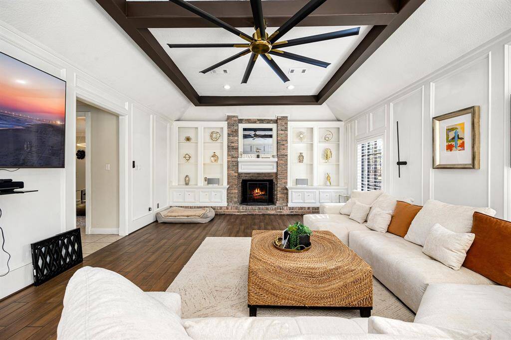 This inviting living room features a cozy brick fireplace, built-in shelving, and a vaulted ceiling with dark wood beams. The space is elegantly furnished with a sectional sofa and woven coffee table, creating a warm ambiance. Large windows flood the room with natural light.