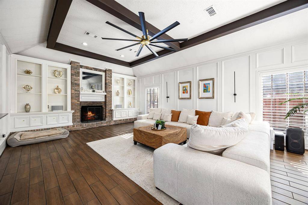 Discover this inviting living room featuring a cozy fireplace, built-in shelving, and a spacious sectional couch. Enjoy the elegance of wood flooring, a modern ceiling fan, and abundant natural light creating a bright, airy atmosphere.