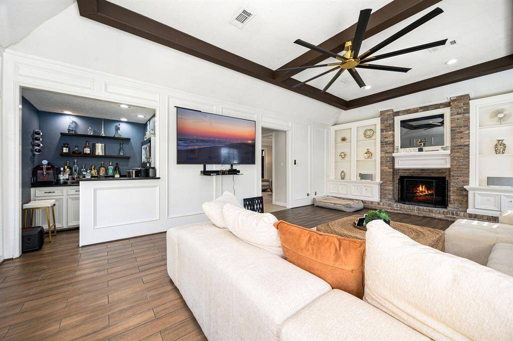 This inviting living room features a cozy stone fireplace, built-in shelves, and a large wall-mounted TV. A sleek wet bar enhances the space, complemented by a modern ceiling fan and plush sectional on stylish wood flooring. Perfect for entertaining!