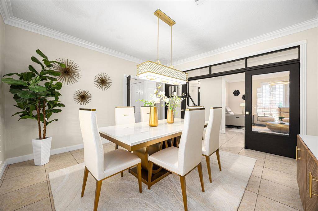 This photo showcases a modern dining room with a sleek white table and high-back chairs. It features a stylish chandelier, decorative wall accents, and a large potted plant. The room is bright with natural light and has a sophisticated, contemporary design.