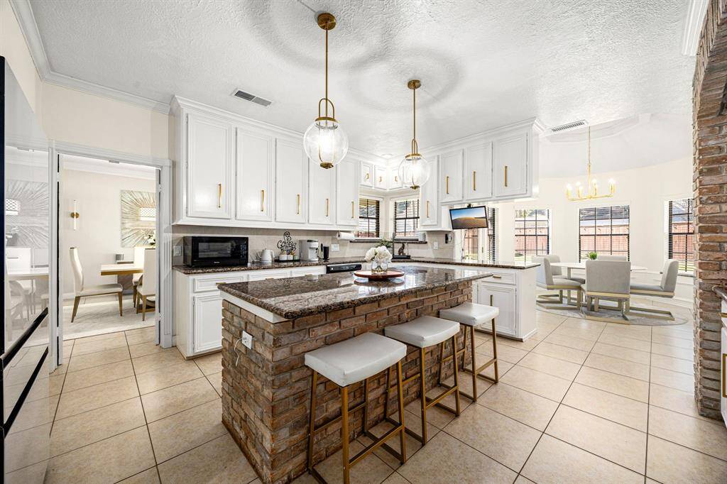 Experience this bright kitchen with elegant white cabinetry, gold handles, a brick island topped with granite, and sleek pendant lighting. It seamlessly connects to a sunlit dining area, creating a warm and inviting space.