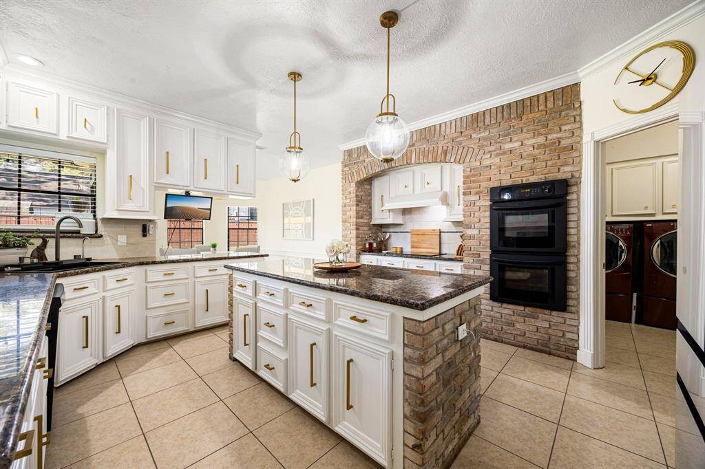 Discover a sleek kitchen with white cabinetry, granite countertops, and a central island. Enjoy the convenience of double wall ovens, a charming brick accent wall, and elegant pendant lighting. The adjacent laundry room is easily accessible through an open doorway.