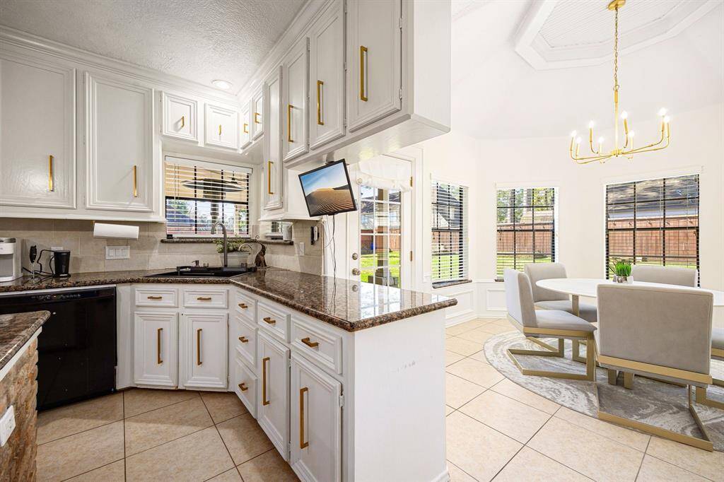 This bright kitchen and dining area boasts elegant white cabinetry with gold handles, granite countertops, and a built-in sink. Enjoy a cozy dining experience with a round table, cushioned chairs, and large windows that flood the space with natural light. A modern chandelier adds a touch of sophistication.