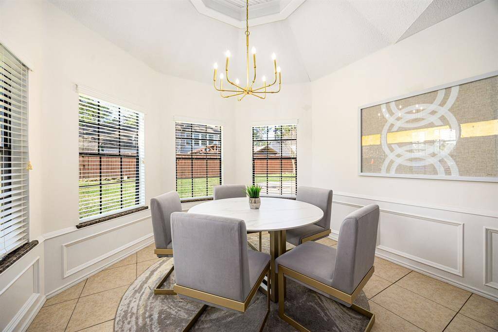 This bright dining area boasts a modern round table with elegant gray chairs, enhanced by large windows for abundant natural light. A contemporary chandelier and tasteful wall art complete the stylish, inviting space.
