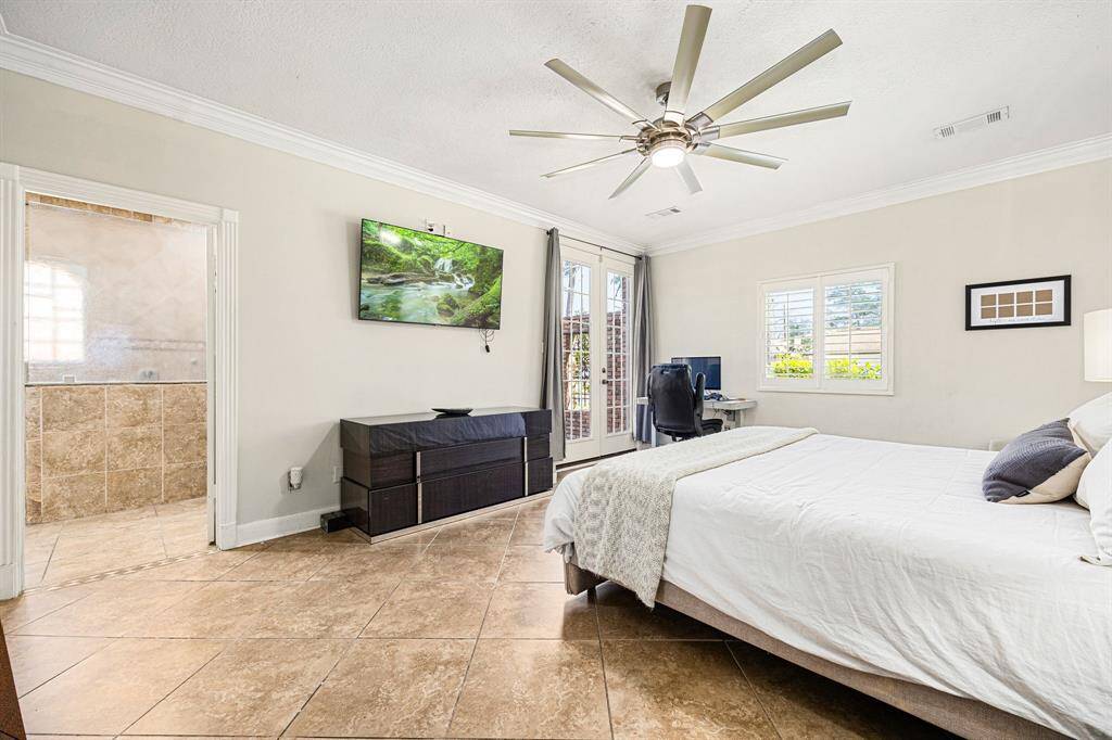 Experience this bright, spacious bedroom featuring tile flooring, a ceiling fan, and French doors to the outside. Enjoy the convenience of an ensuite bathroom with neutral tiles, a wall-mounted TV, and a cozy office nook by the window.