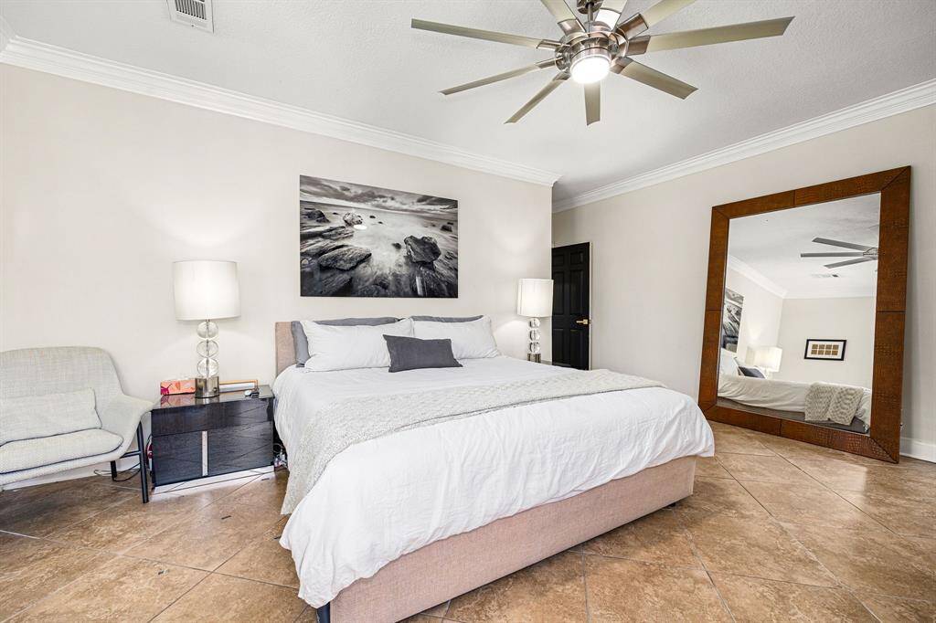 This inviting bedroom boasts a modern ceiling fan, elegant tile flooring, and neutral tones. It features a large bed, stylish side tables with lamps, a cozy chair, and a striking floor mirror.