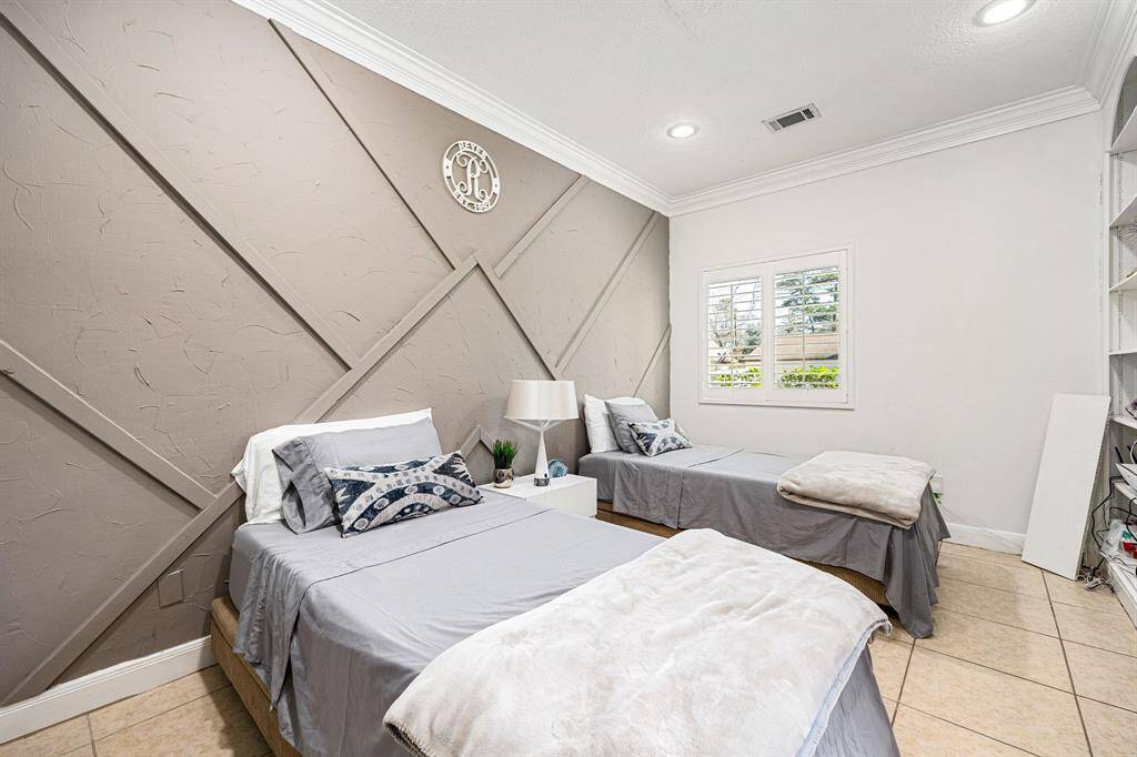 Experience comfort in this stylish bedroom with a geometric accent wall, two twin beds, and elegant gray bedding. Natural light flows through plantation shutters, complemented by recessed lighting, tiled flooring, and built-in shelving for added storage.