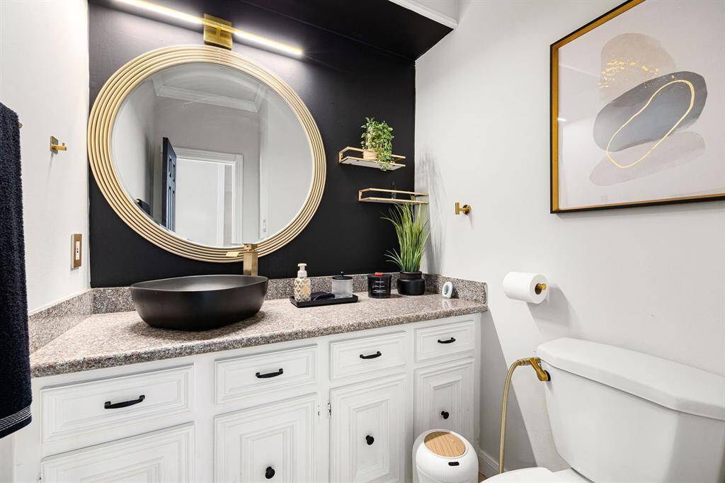 This stylish bathroom boasts a modern black accent wall, a striking gold-framed mirror, and a sleek black vessel sink. Elegant white cabinetry offers ample storage, complemented by decorative plants, contemporary artwork, and gold fixtures.