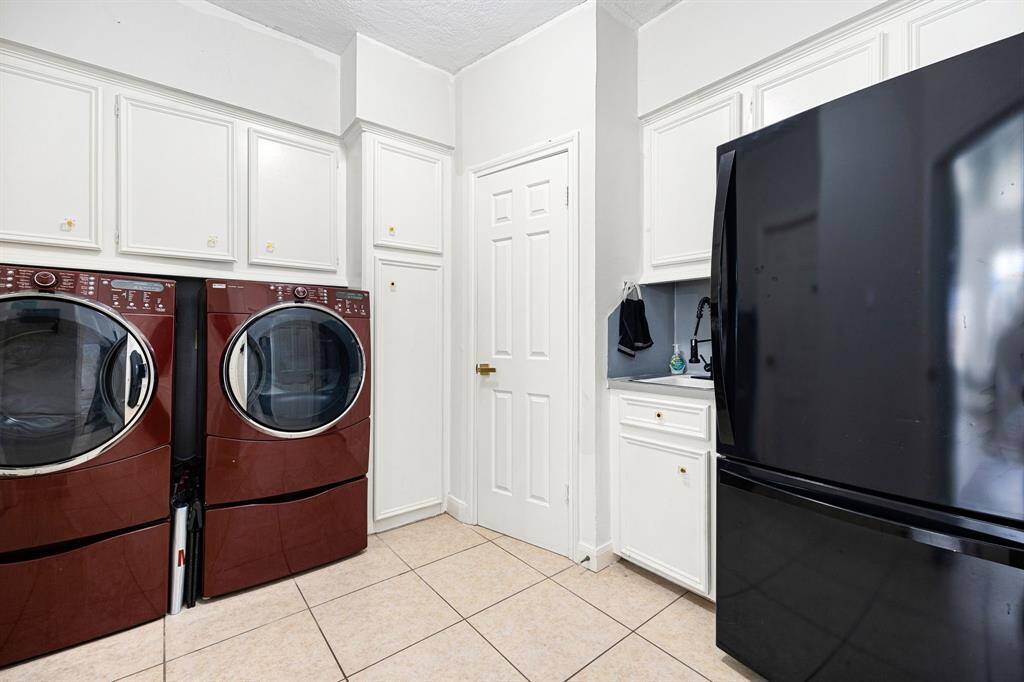 Discover a stylish laundry room featuring sleek, dark red front-loading washer and dryer units. Enjoy ample white cabinetry, a modern black refrigerator, and clean tiled flooring for a functional and elegant space.