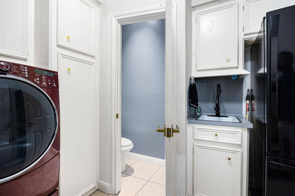 This efficient laundry area features a sleek front-load washer and dryer, ample white cabinetry for storage, and a modern sink with a black faucet. Conveniently located next to a bathroom, it also includes a dark refrigerator for added utility.