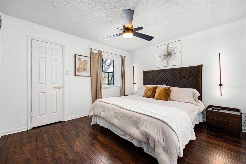 This modern bedroom offers a cozy retreat with a comfortable bed, elegant headboard, and sleek ceiling fan. Enjoy the warmth of hardwood floors, neutral walls, and abundant natural light for an inviting atmosphere.