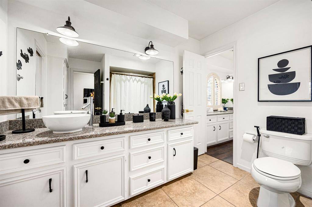 This bathroom features a modern design with a large mirror, stylish vessel sink, and granite countertop. The white cabinetry provides ample storage, and the decor includes contemporary black accents and artwork. Natural light enhances the clean, bright atmosphere.