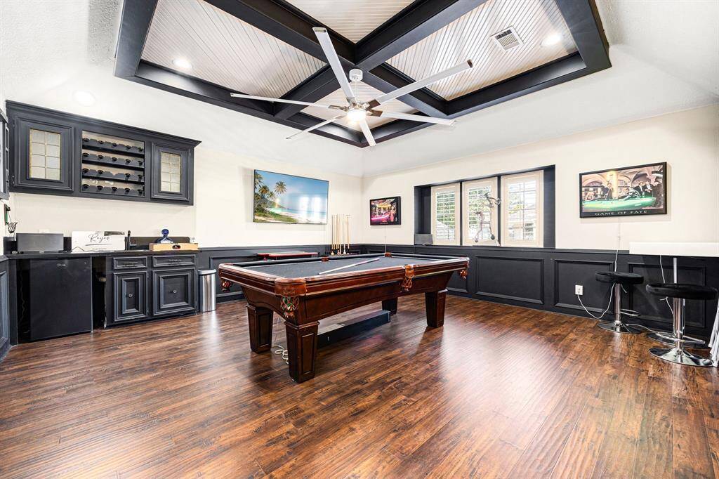 Discover the ultimate entertainment space with this expansive game room, featuring a stylish pool table, elegant dark wood flooring, and a stunning coffered ceiling with a large fan. The built-in bar with cabinetry and seating is perfect for hosting, while large windows flood the room with natural light.