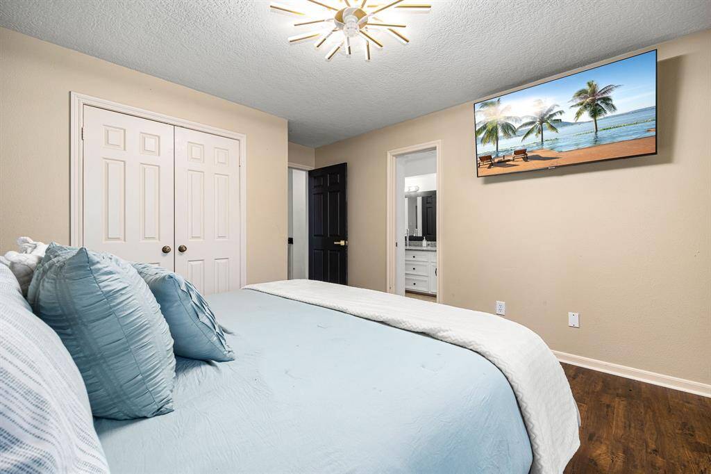 Experience comfort in this inviting bedroom featuring warm beige walls, elegant wood flooring, and a soft blue bedspread. Enjoy modern amenities with a stylish light fixture, wall-mounted TV, and a spacious double-door closet. The en-suite bathroom offers added convenience and a chic vanity view.