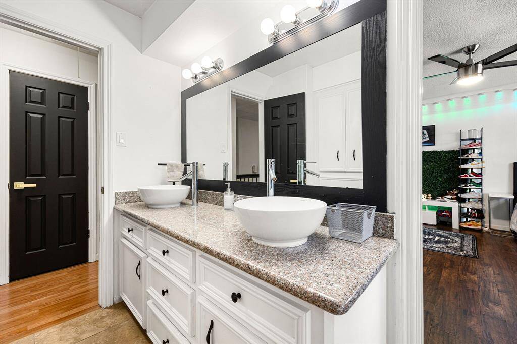 Experience luxury in this modern bathroom featuring dual vessel sinks on a granite countertop, complemented by white cabinets and a large mirror with stylish lighting. The adjacent room boasts elegant wood flooring, a ceiling fan, and a chic shoe display rack.