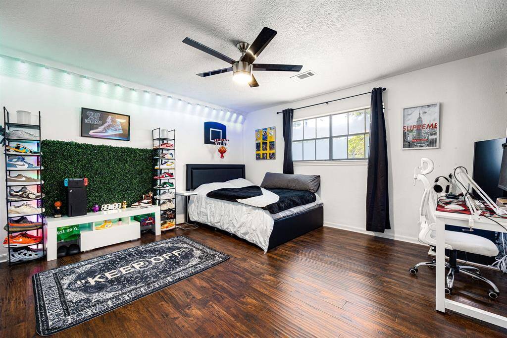 Discover a stylish bedroom featuring hardwood floors, a modern ceiling fan, and abundant natural light. This space includes a cozy bed, a functional desk, and ample shelving. Accented with a shoe display, sports memorabilia, and ambient LED lighting, it's the perfect blend of style and functionality.