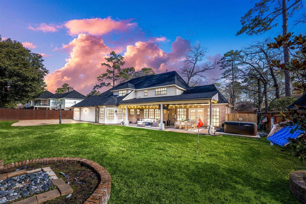 Discover this charming home with a spacious backyard, featuring a lush lawn, cozy covered patio, and relaxing hot tub. Large windows and elegant lighting enhance its appeal, set against mature trees and a stunning sunset sky.