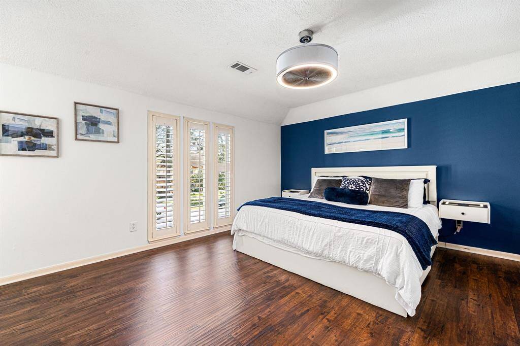 This inviting bedroom boasts rich hardwood floors, a striking blue accent wall, and abundant natural light. It features a large bed, contemporary side tables, and a modern light fixture, creating a warm and cozy atmosphere.