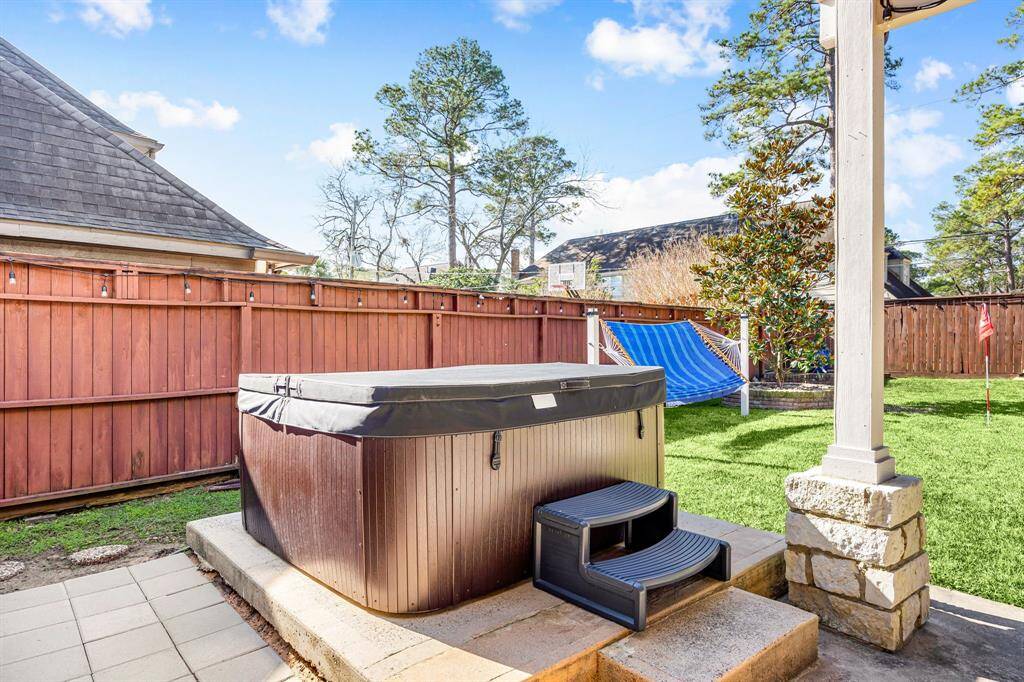 Discover your serene outdoor retreat with a cozy hot tub on a paved patio, nestled in a lush green lawn. Enjoy privacy with a wooden fence and unwind in a blue hammock. Mature trees and a charming stone pillar complete this tranquil setting.