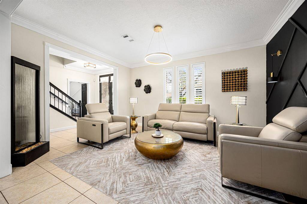 Experience modern elegance in this living room, featuring sleek leather seating, a geometric accent wall, and a contemporary chandelier. Sunlight floods through large windows, enhancing the ambiance with a unique water feature.