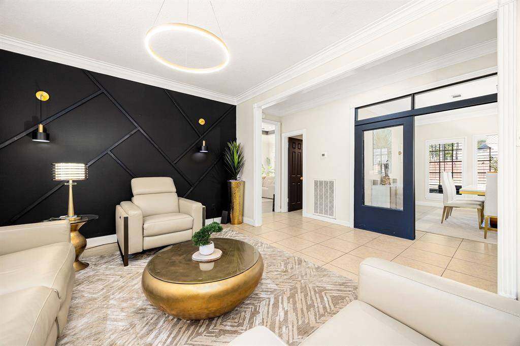 Experience modern elegance in this living area, featuring a striking black accent wall with geometric patterns and sleek lighting. Enjoy the comfort of beige seating, a chic gold coffee table, and a bright, tiled floor. A wide doorway with dark trim connects to an adjacent space, inviting abundant natural light.
