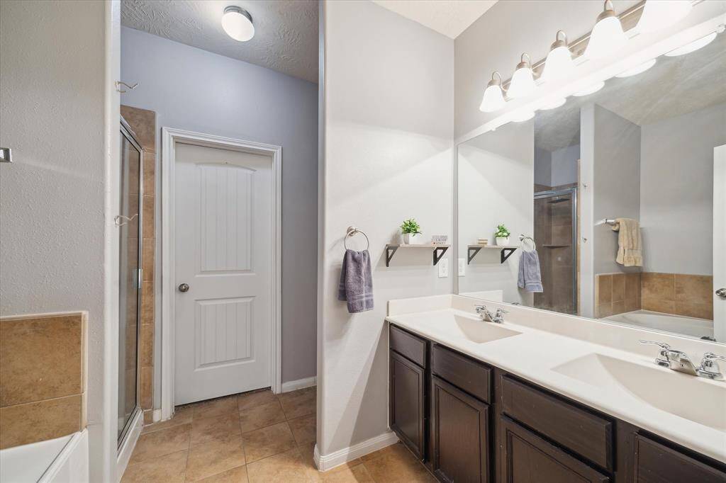 En suite primary bath features a double vanity with dark wood cabinets and a large mirror. It includes a walk-in shower, neutral tile flooring, and modern lighting. Small shelves add extra storage and decor space.