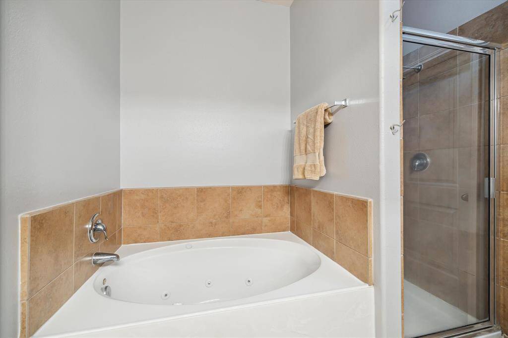 Priimary bath features a large corner jetted tub with tan tile accents, a modern glass-enclosed shower, and neutral wall colors, creating a relaxing and elegant space.