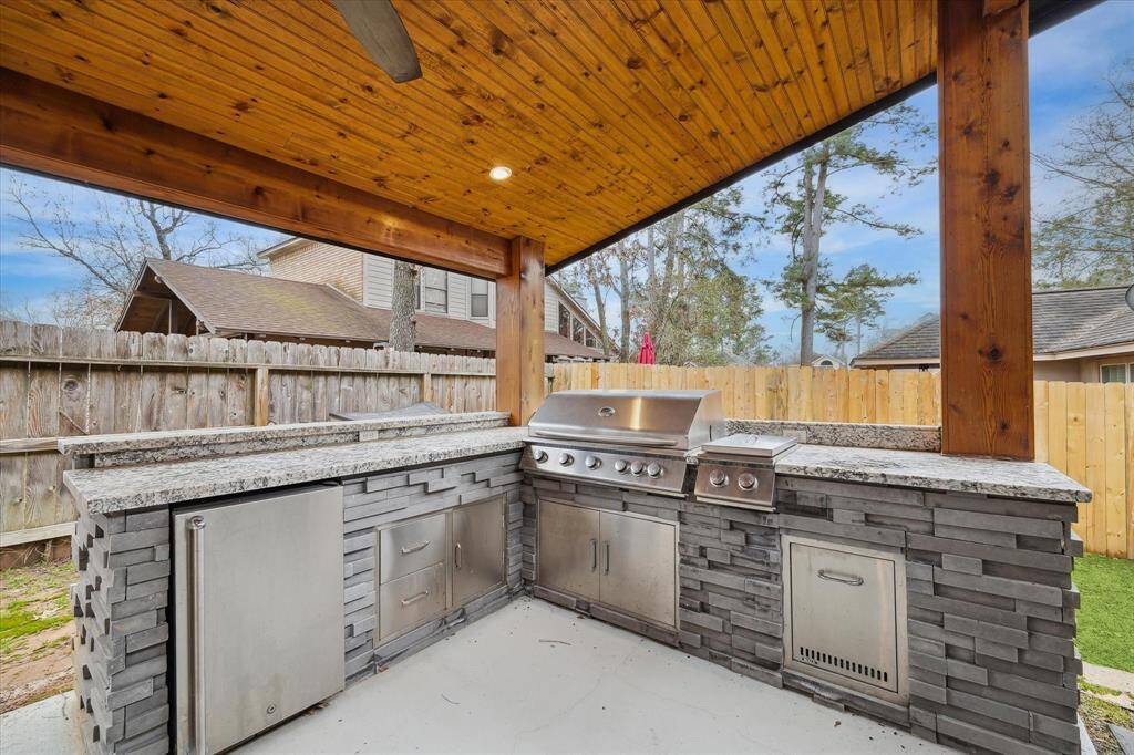 This photo showcases a stylish outdoor kitchen with a brick and stone design, featuring a refrigerator, built-in grill and ample counter space under a wooden pergola. Perfect for entertaining in a private backyard setting.