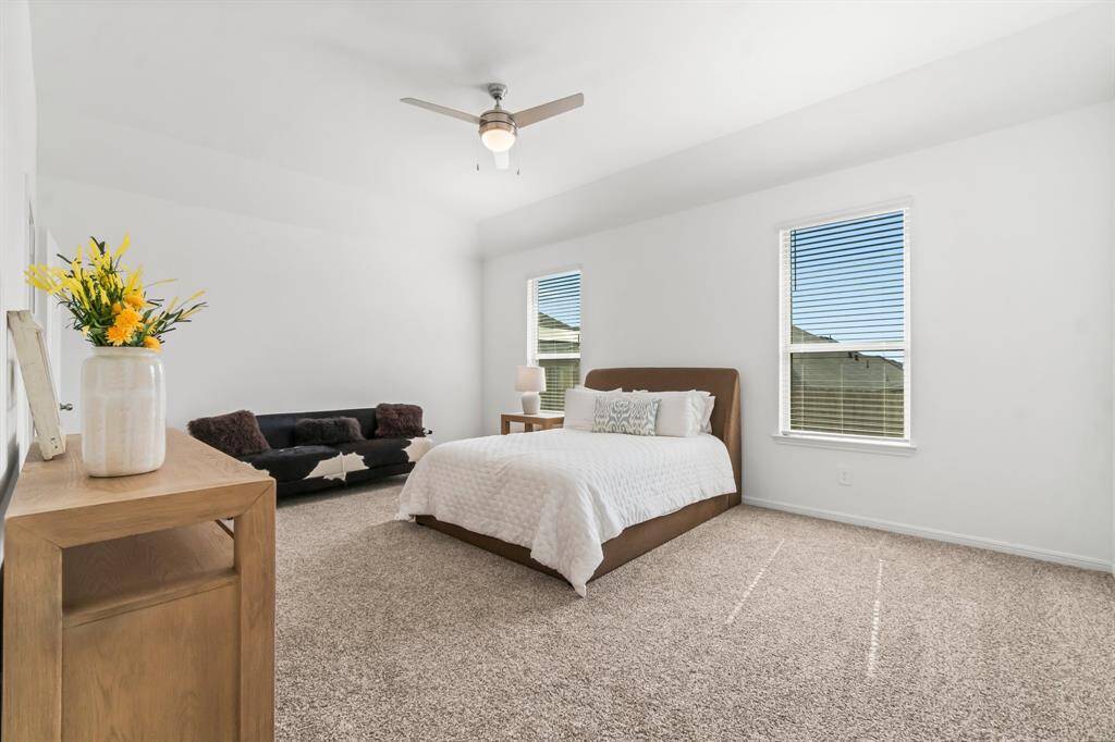 This is a bright and spacious primary bedroom with plush carpeting, large windows for natural light, a ceiling fan for comfort, and a cozy seating area. The neutral color palette offers a serene and adaptable space for relaxation.