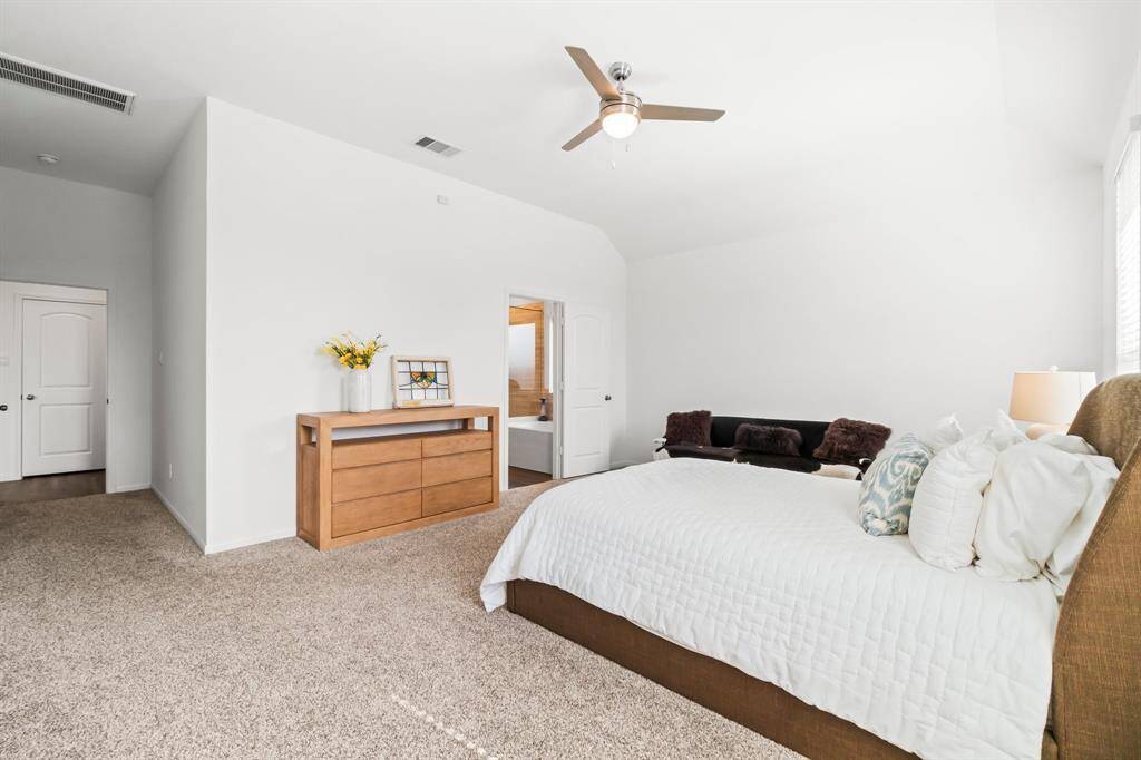 Spacious, well-lit bedroom with neutral walls, carpeted flooring, high ceiling with a fan, and large windows.