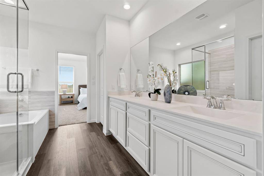 This is a bright and modern ensuite bathroom featuring a double vanity with ample storage, a glass-enclosed shower, and a view into the adjoining primary bedroom. The space is accented with contemporary finishes and a neutral color palette.