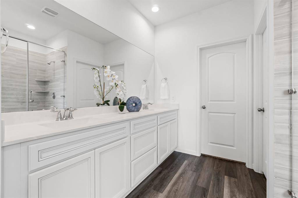 Modern, bright primary bathroom featuring a double vanity with ample storage, elegant marble-like tiling, a glass-enclosed shower, and stylish hardwood floors. There are two large separate walk-in closets.