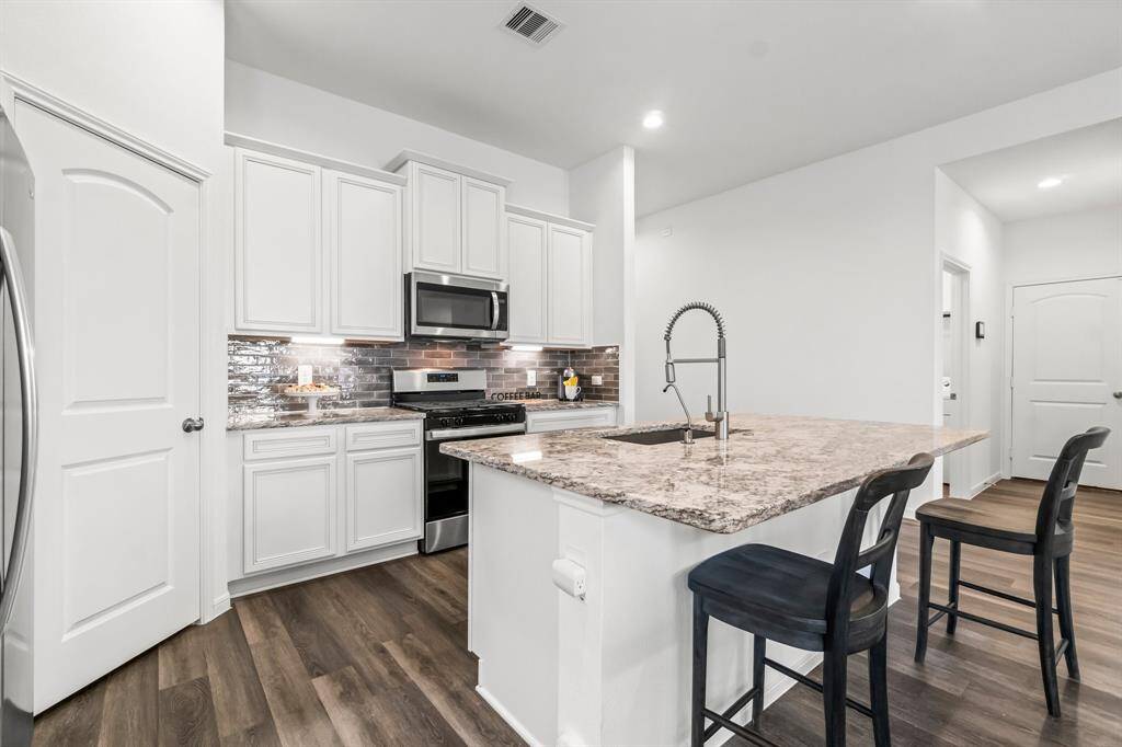 This is a modern kitchen featuring white cabinetry, stainless steel appliances, a mosaic tile backsplash, granite countertops, and a central island with seating space.