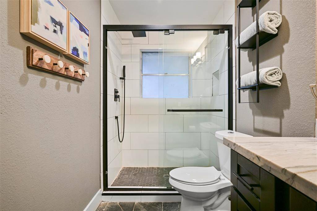 The master bathroom has been beautifully renovated to include a sleek black vanity with a granite top and a rectangular basin sink, a circular framed mirror, contemporary lighting, slate tile floors, and a glass door shower with dual showerheads and a tile surround.