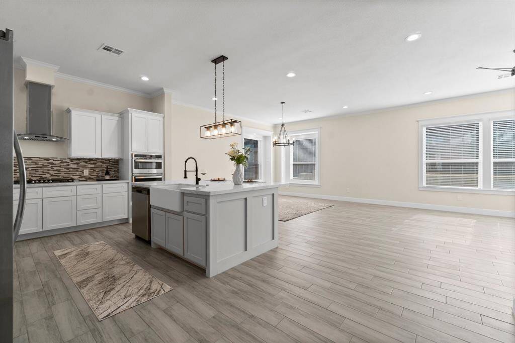This photo showcases a spacious, open-concept kitchen and living area. The kitchen features modern white cabinetry, stainless steel appliances, and a central island with a farmhouse sink. The space is bright, with ample natural light from large windows and stylish light fixtures. The flooring is a sleek, light wood finish, enhancing the contemporary feel.