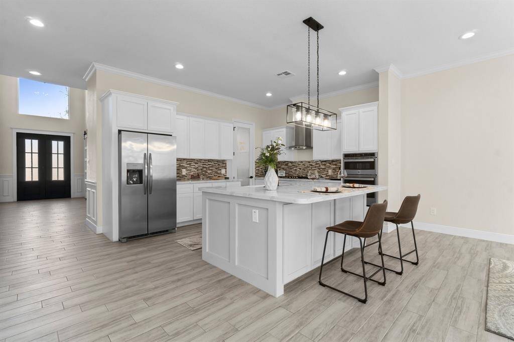 This modern kitchen features sleek white cabinetry, a large island with seating, stainless steel appliances, and a stylish tile backsplash. The space is open and bright with recessed lighting and light wood flooring, offering a welcoming atmosphere.
