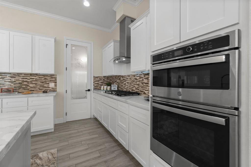 This modern kitchen features sleek white cabinetry, stainless steel double ovens, a tiled backsplash, and wood-look flooring. It offers a clean, contemporary design with ample counter space and a convenient pantry door.