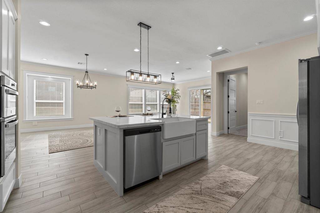 This photo showcases a modern, open-concept kitchen and dining area with elegant light fixtures, a central island with a sink, stainless steel appliances, and light wood flooring. Large windows provide ample natural light, creating a bright and inviting space.