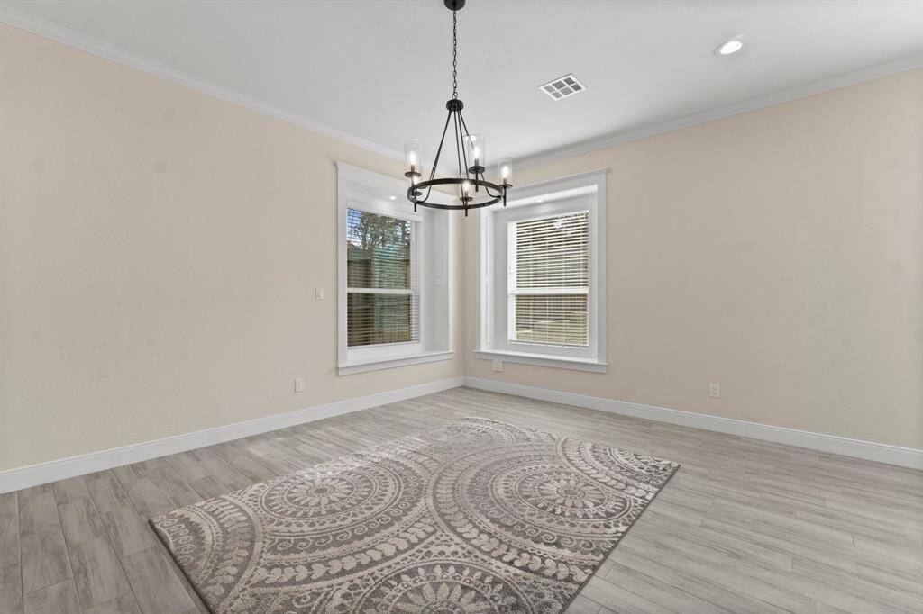This room features light hardwood flooring, a decorative area rug, and neutral walls. Two large windows provide natural light, and a modern chandelier adds elegance. Ideal for a dining or sitting area.