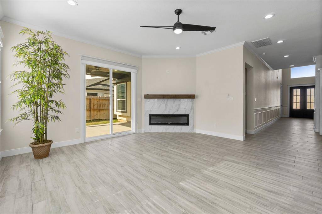 This spacious, modern living area features light wood-look flooring, a stylish ceiling fan, and a sleek built-in mock fireplace. Large sliding glass doors lead to a backyard, enhancing the room with natural light.