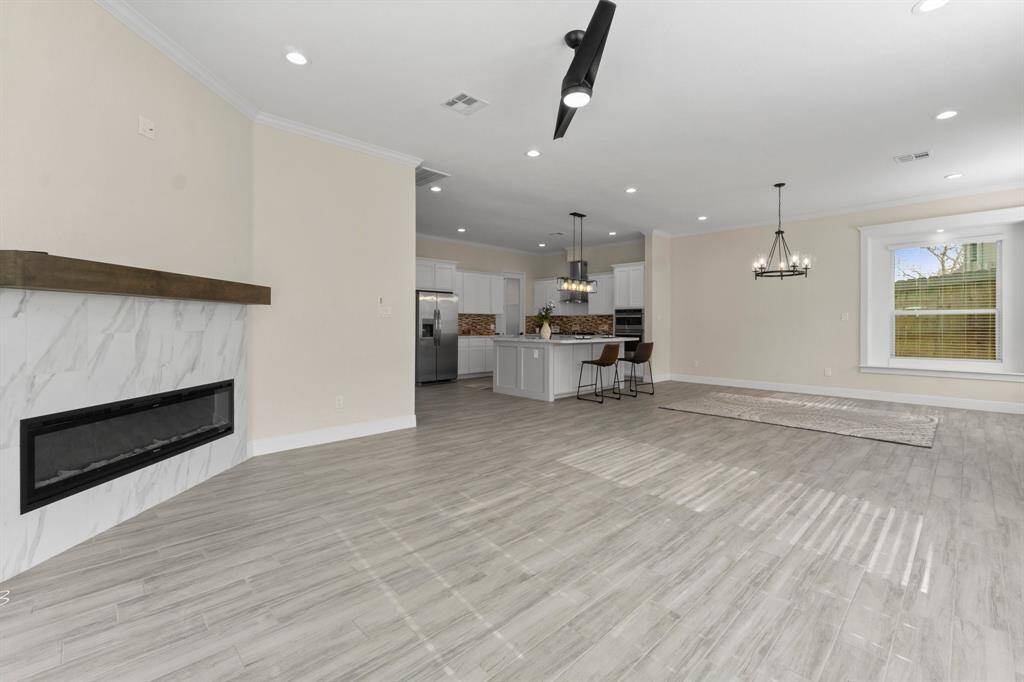 This spacious, open-concept living area features modern wood-look flooring, a sleek built-in fireplace, and a contemporary kitchen with a large island and stainless steel appliances. Ample natural light fills the room, highlighting the modern light fixtures and neutral color palette.