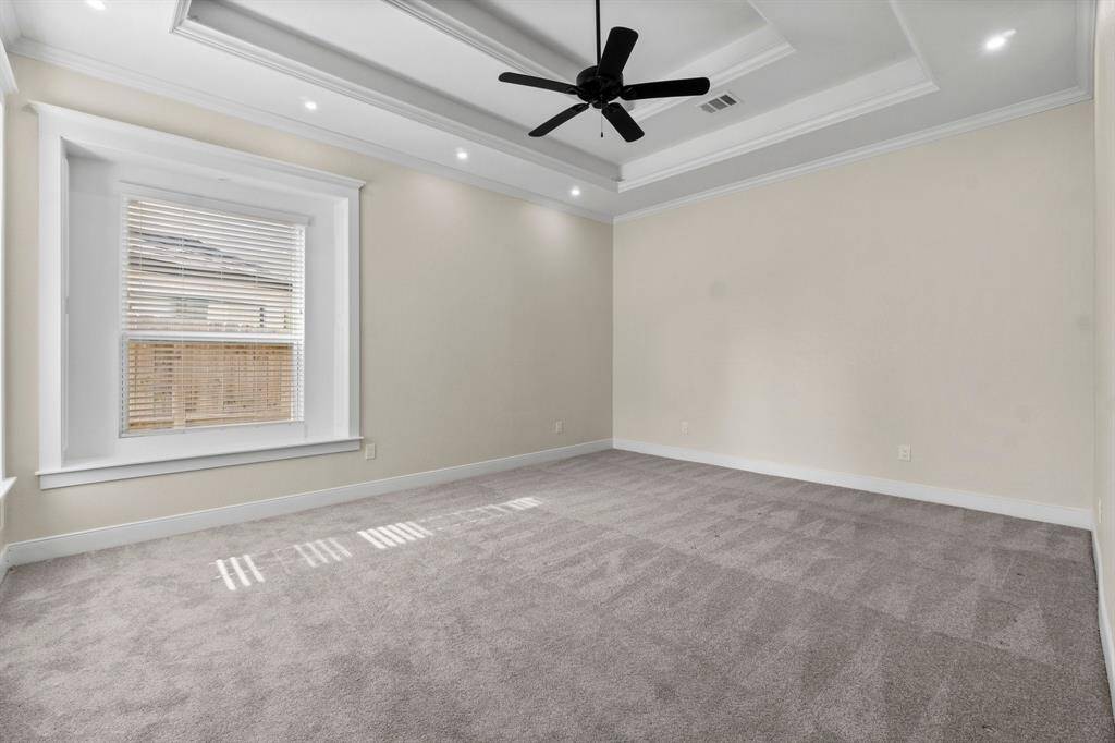This spacious primary bedroom features neutral walls, plush carpeting, a large window for natural light, and a stylish tray ceiling with recessed lighting and a ceiling fan.