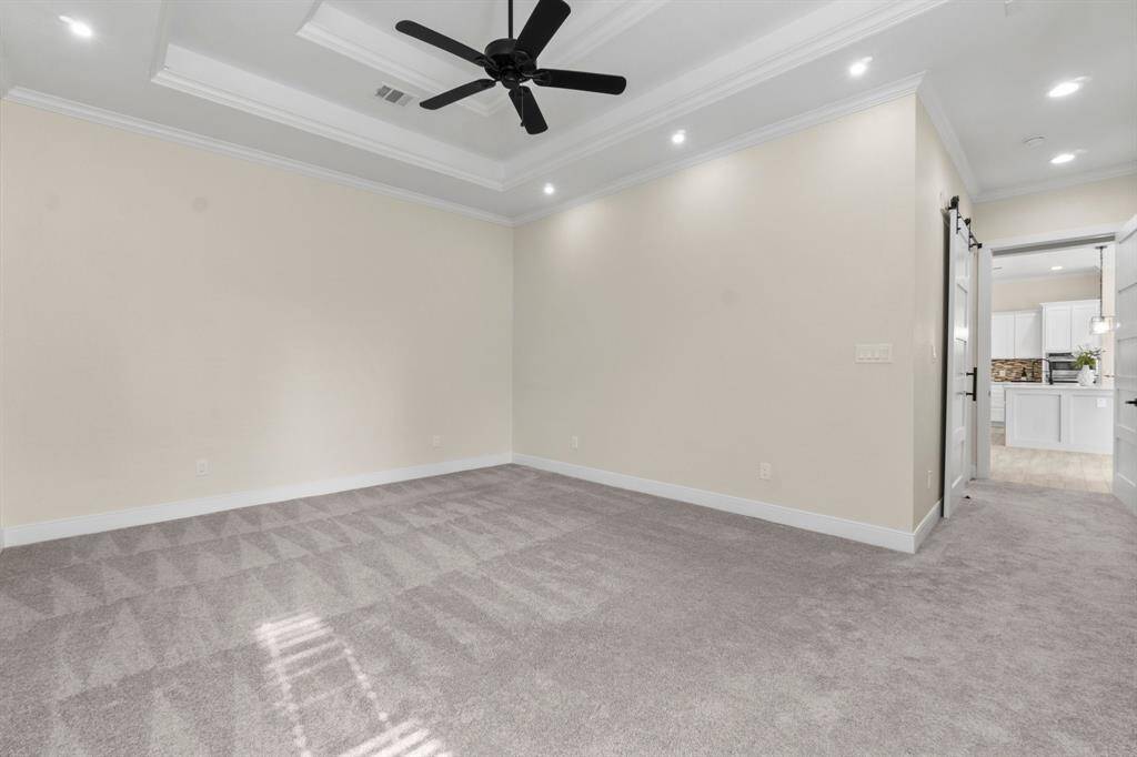 This primary bedroom features soft beige walls and plush carpeting, accented by a tray ceiling with recessed lighting and a modern black ceiling fan.
