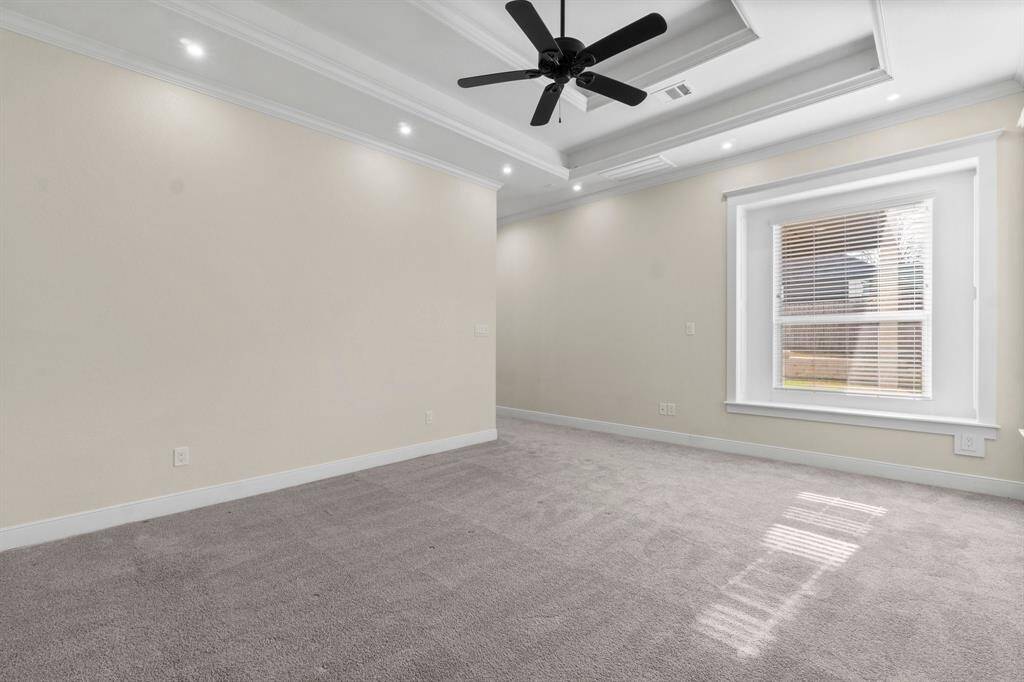 This primary bedroom features soft carpeted flooring, a tray ceiling with recessed lighting, and a ceiling fan. A large window allows natural light to fill the space, creating a bright and inviting atmosphere.