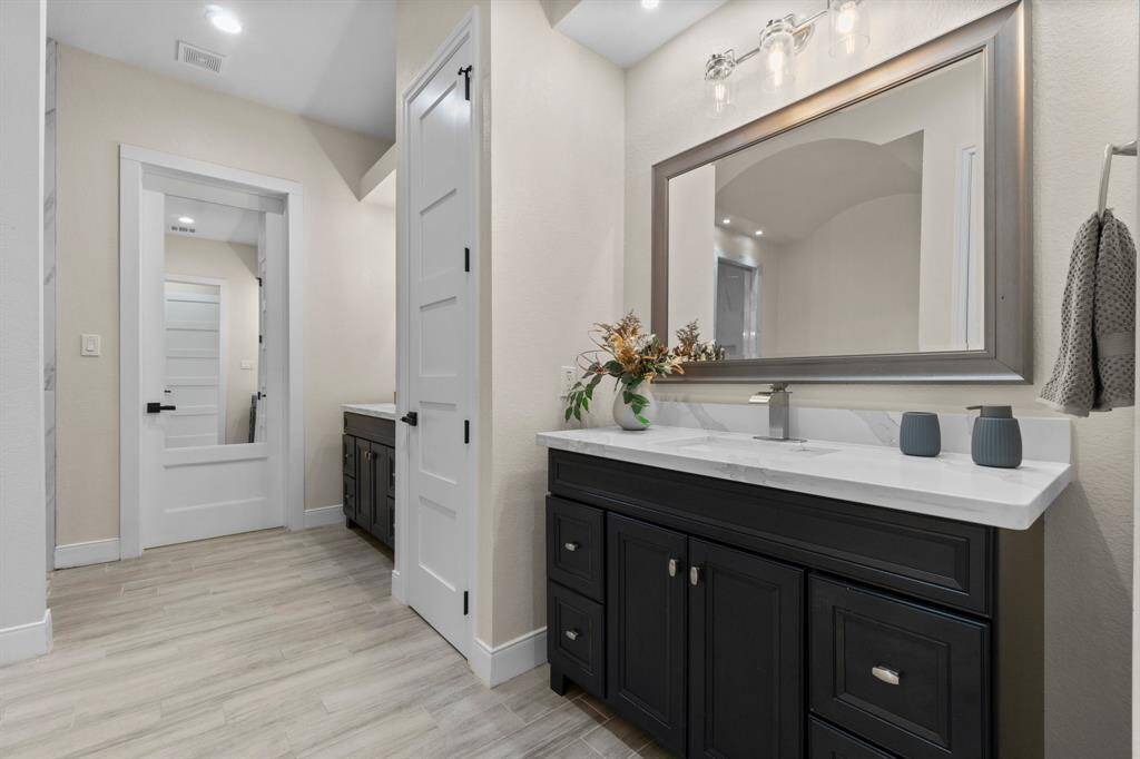 This photo showcases a modern primary bathroom with dual sinks, dark cabinetry, and sleek fixtures. It features a large mirror, light walls, and a wood-look floor, creating a clean and inviting space.