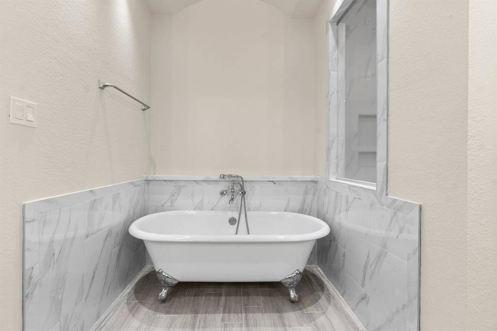 The primary bathroom features a classic clawfoot tub with stylish marble-patterned wall tiles and light wood-look flooring, creating a clean and elegant space.