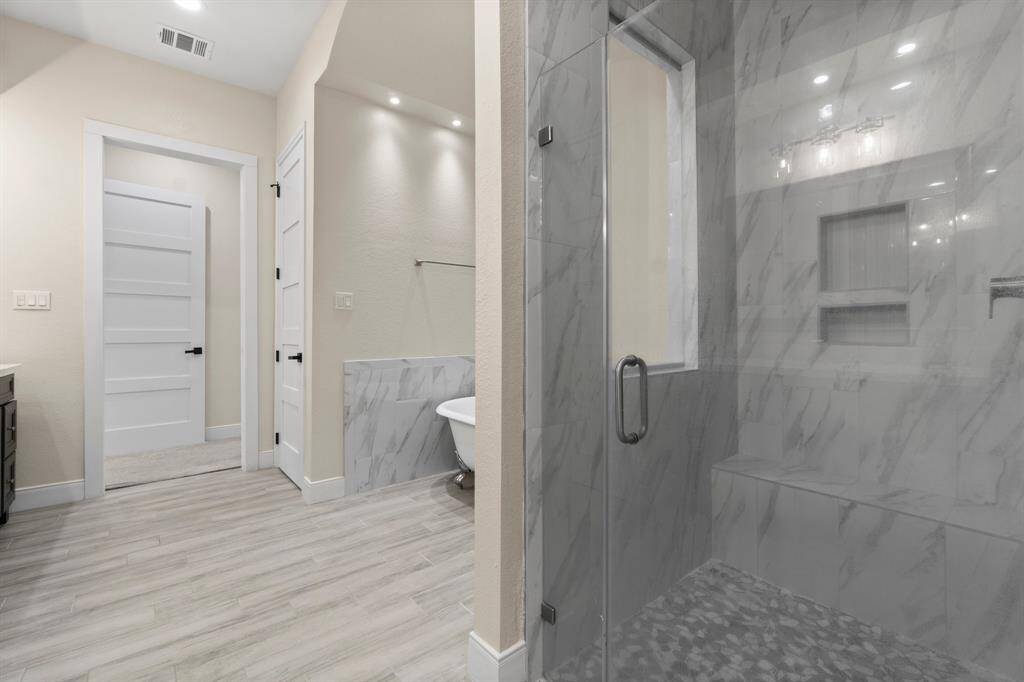 This modern primary bathroom features a sleek glass-enclosed shower with elegant marble-style tiles. The room includes a freestanding bathtub, light wood-style flooring, and recessed lighting, creating a bright and inviting space.
