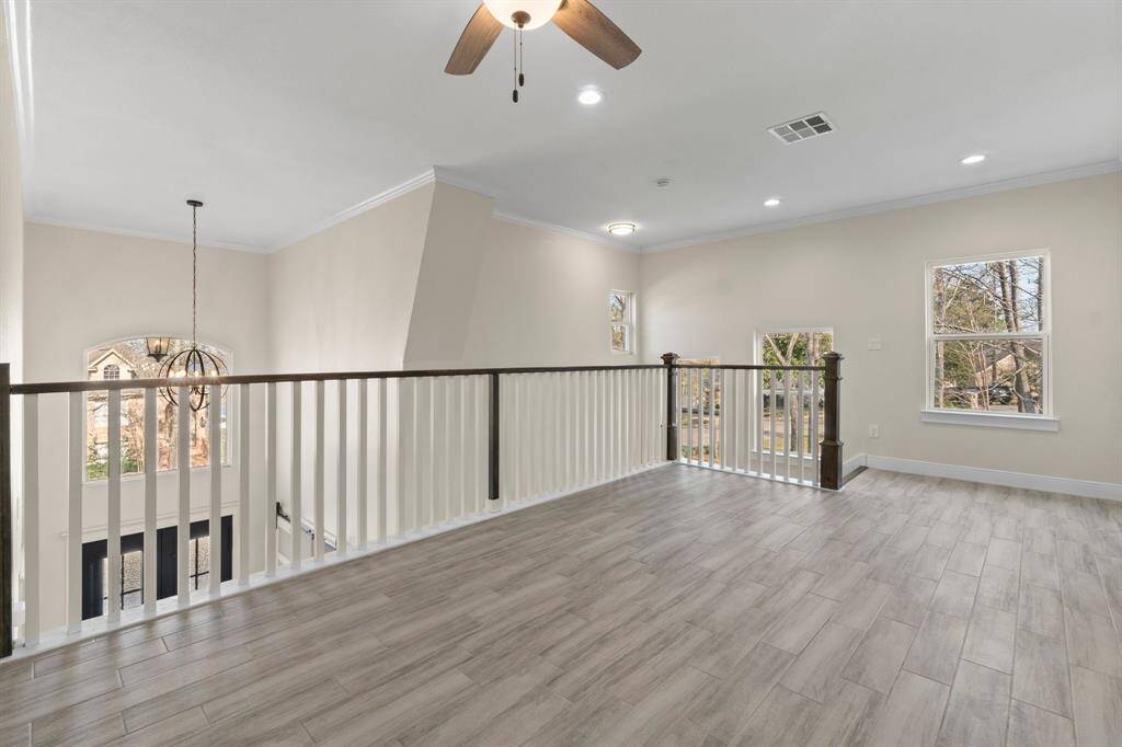 This photo showcases a bright, open loft area with light wood flooring and neutral walls. It features a ceiling fan, recessed lighting, and a view of the outdoors through large windows. The space overlooks a foyer with a modern chandelier, offering a spacious and airy feel.