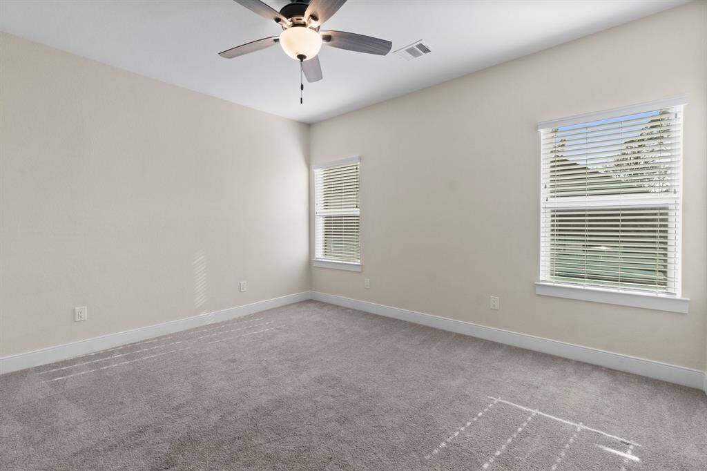 This photo shows a bright, spacious room with neutral walls and carpeted flooring. It features a ceiling fan and two windows with blinds, allowing plenty of natural light.